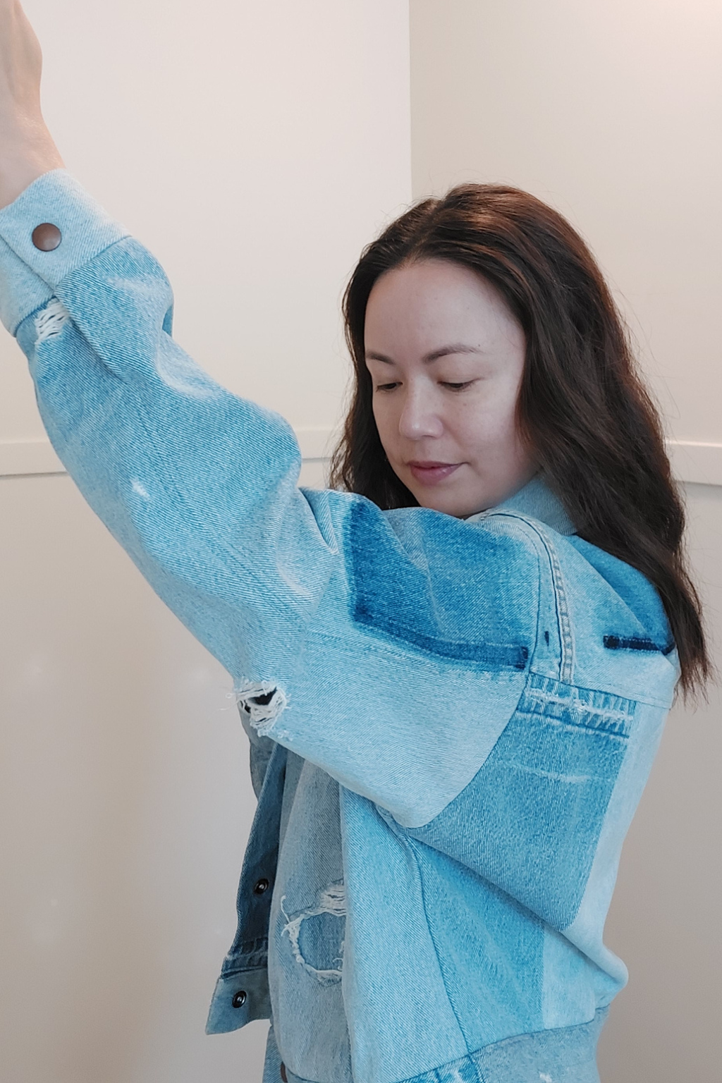 A woman wearing a light blue patchwork denim jacket, with one arm raised, standing against a plain off-white wall. Upcycled denim jacket by September Archive. Photo by Sheila Wong.
