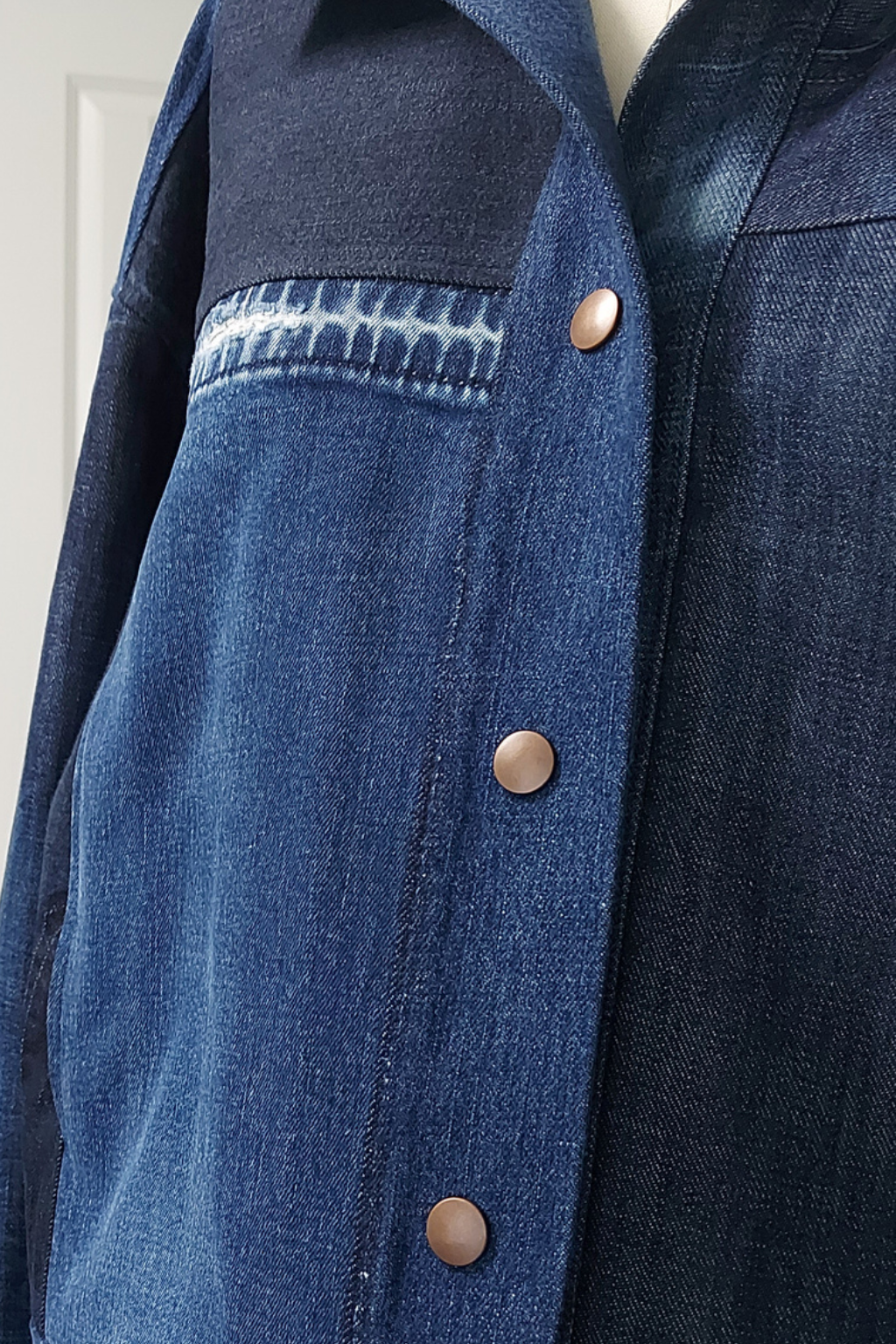 Close-up of a dark blue denim jacket with a contrasting dark navy collar, a chest pocket with a lighter blue stitched detail, and bronze-colored snap buttons. Upcycled denim jacket by September Archive.