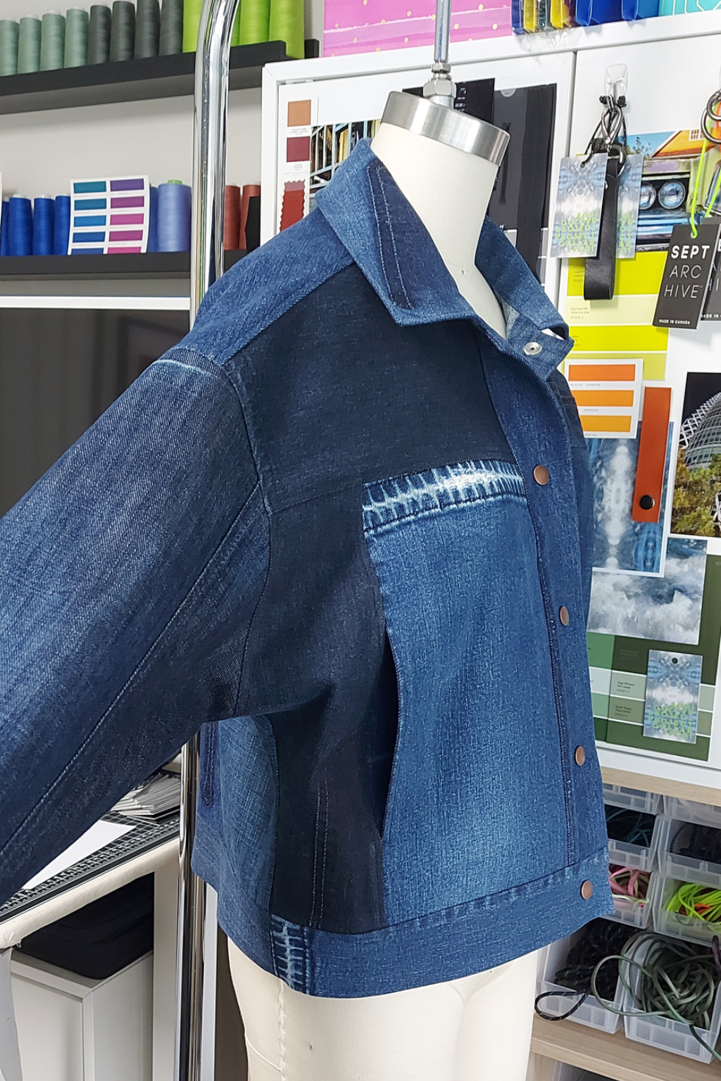 A cropped blue denim jacket with a mixed-panel design, featuring a dark indigo body and lighter blue sleeves and chest pocket, displayed on a mannequin torso in a studio setting.