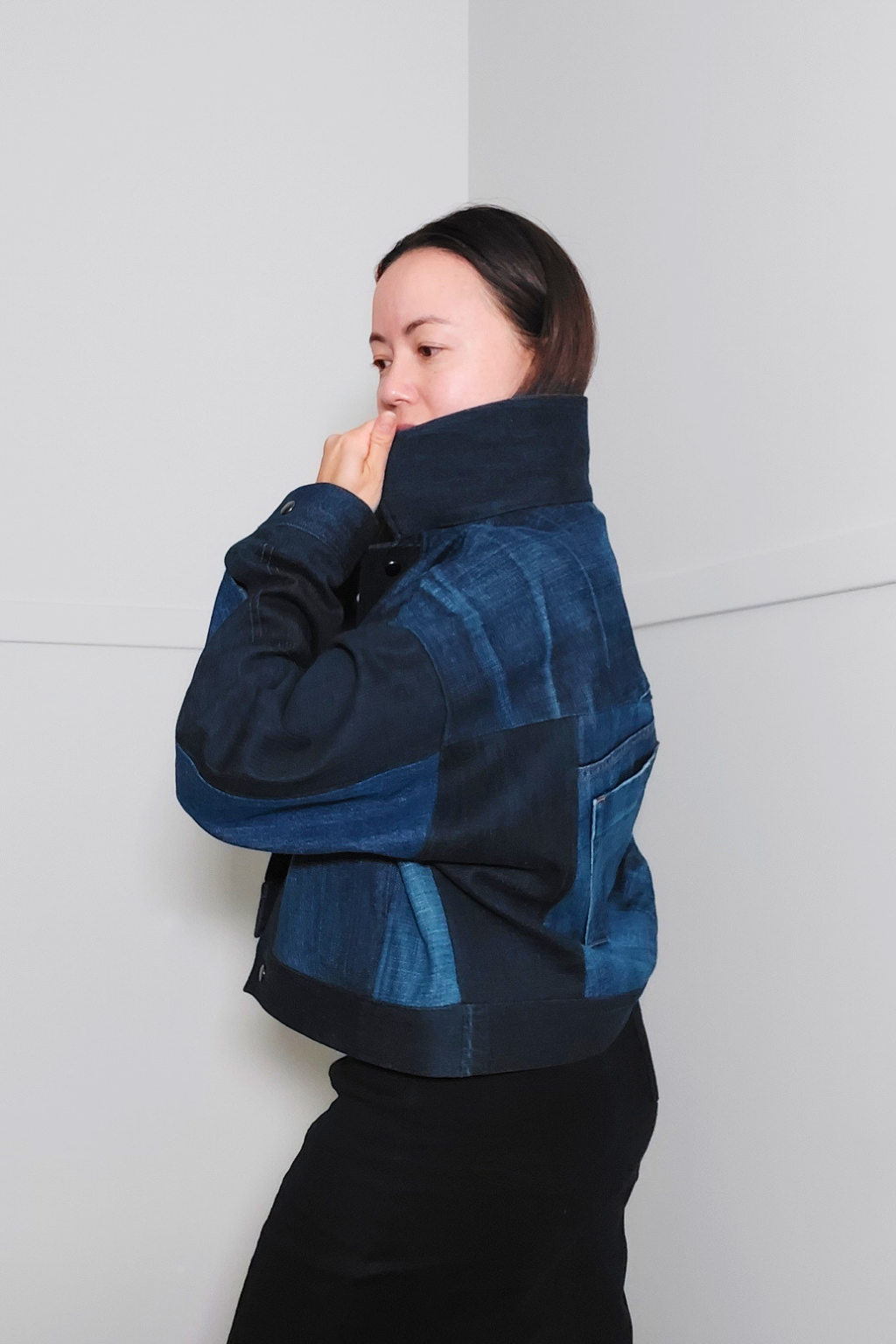 A woman wearing a dark blue patchwork denim jacket with a high collar, posed against a minimalist light gray wall. Upcycled denim jacket by September Archive. Photographed by Sheila Wong.