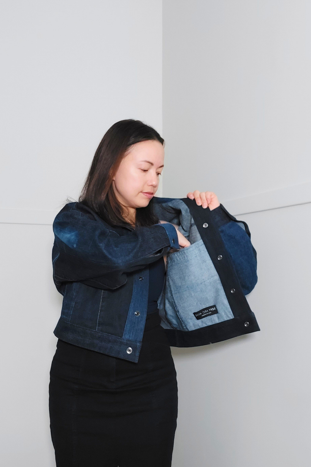 A woman wearing a dark blue patchwork denim jacket and black skirt, holding the jacket open to show the light blue denim lining. The jacket features a dark blue denim front and sleeves with a contrasting light blue denim lining. Upcycled denim jacket by September Archive. Photographed by Sheila Wong.