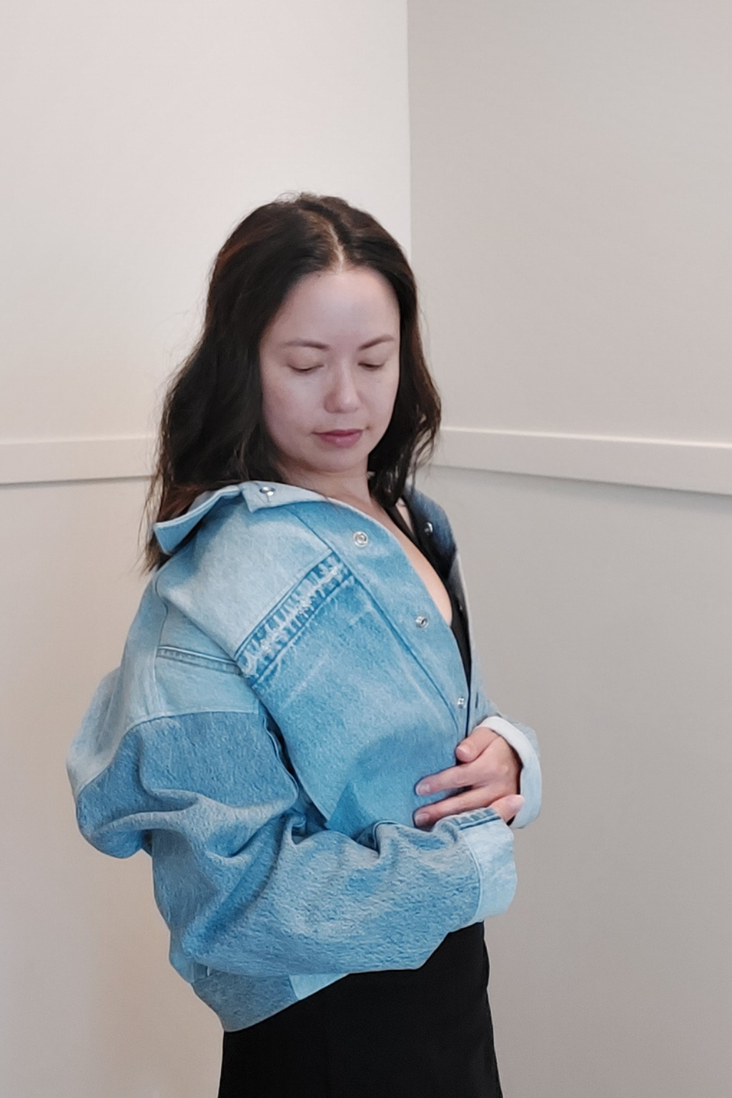 A woman wearing a light blue cropped denim jacket over a black top, standing against a plain off-white wall. Upcycled denim jacket by September Archive. Photo by Sheila Wong.