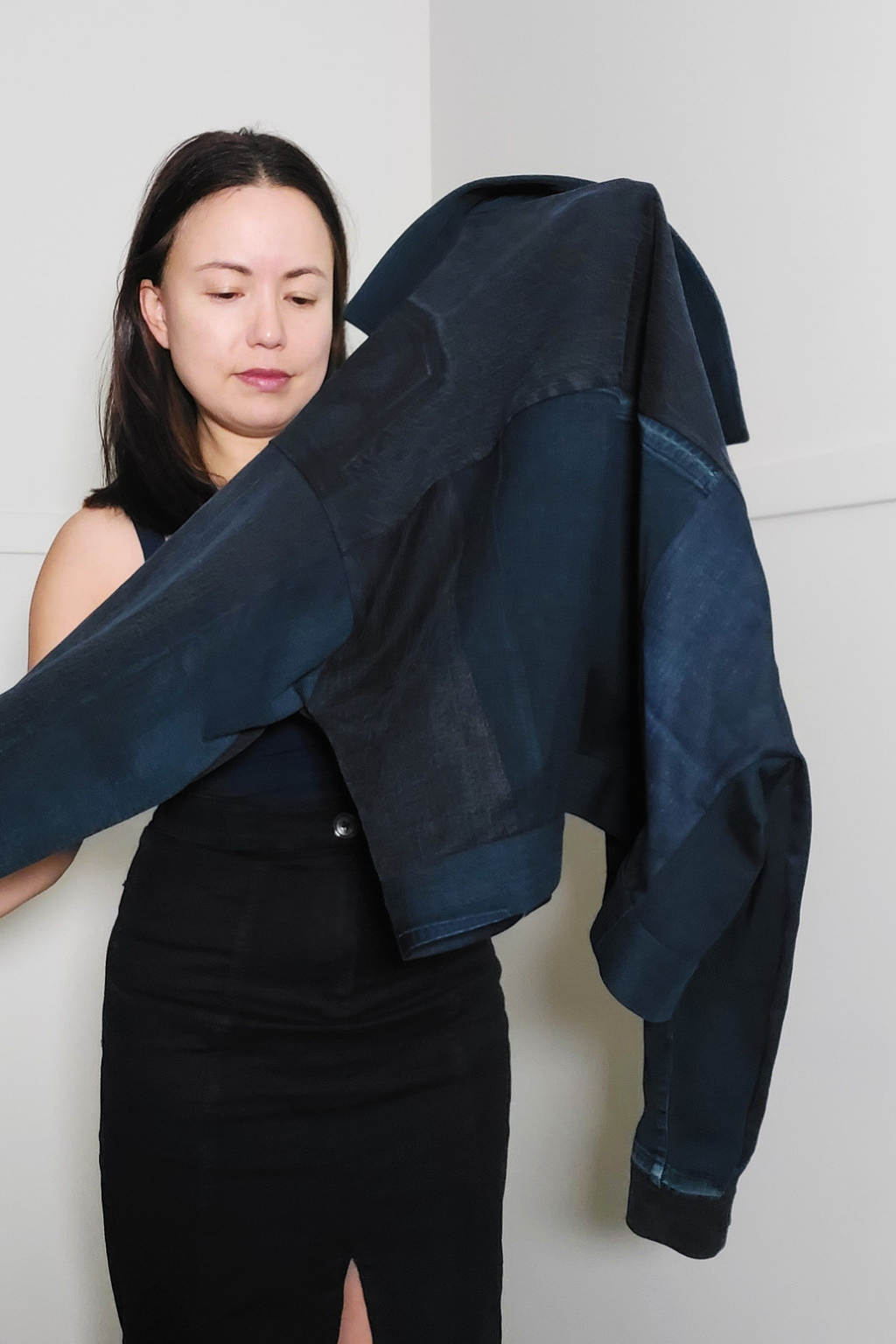 A woman wearing a black dress holds up a dark navy blue cropped jacket made from layered denim panels, set against a plain light gray wall. Upcycled denim jacket by September Archive. Photographed by Sheila Wong.