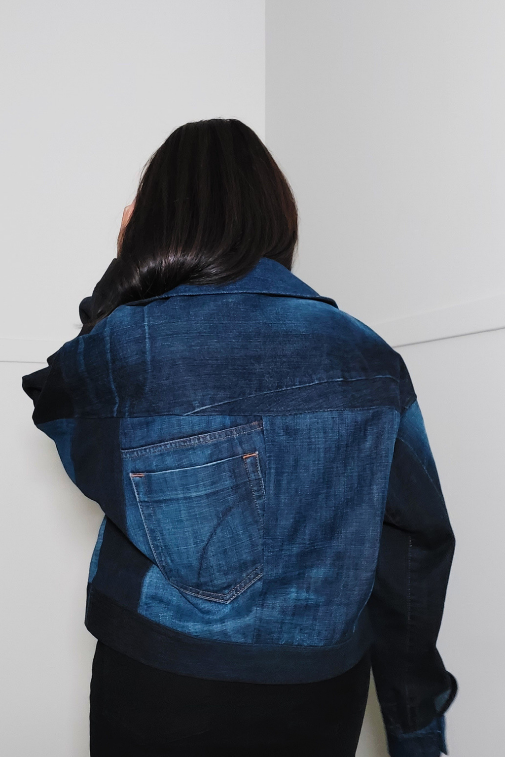 A person viewed from behind wearing a cropped denim jacket with contrasting dark navy sleeves and a back pocket, standing against a plain white wall. Upcycled denim jacket by September Archive. Photographed by Sheila Wong.