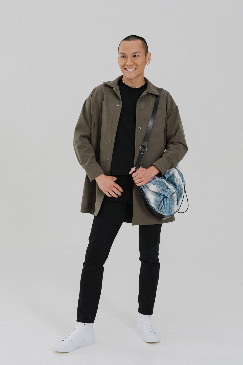 A man modeling an olive green hooded jacket over a black shirt and black pants, carrying a blue and white patterned shoulder bag, against a light gray studio background. Project collab #001: Carryall City Sling bag with collaborative all-over print by September Archive Apparel. Captured by Paper Crane Creative.