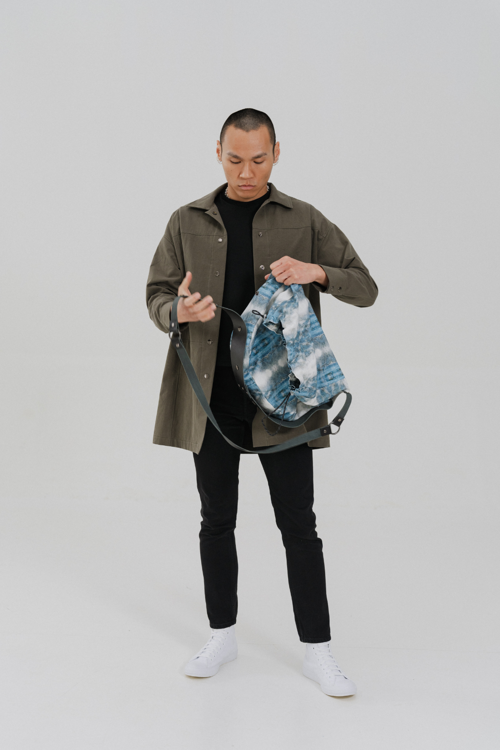 A man wearing a brown jacket, black t-shirt, black pants, and white sneakers is holding a blue and white patterned backpack with a dark strap against a light gray studio background. Project collab #001: Carryall City Sling bag with collaborative all-over print by September Archive Apparel. Captured by Paper Crane Creative.