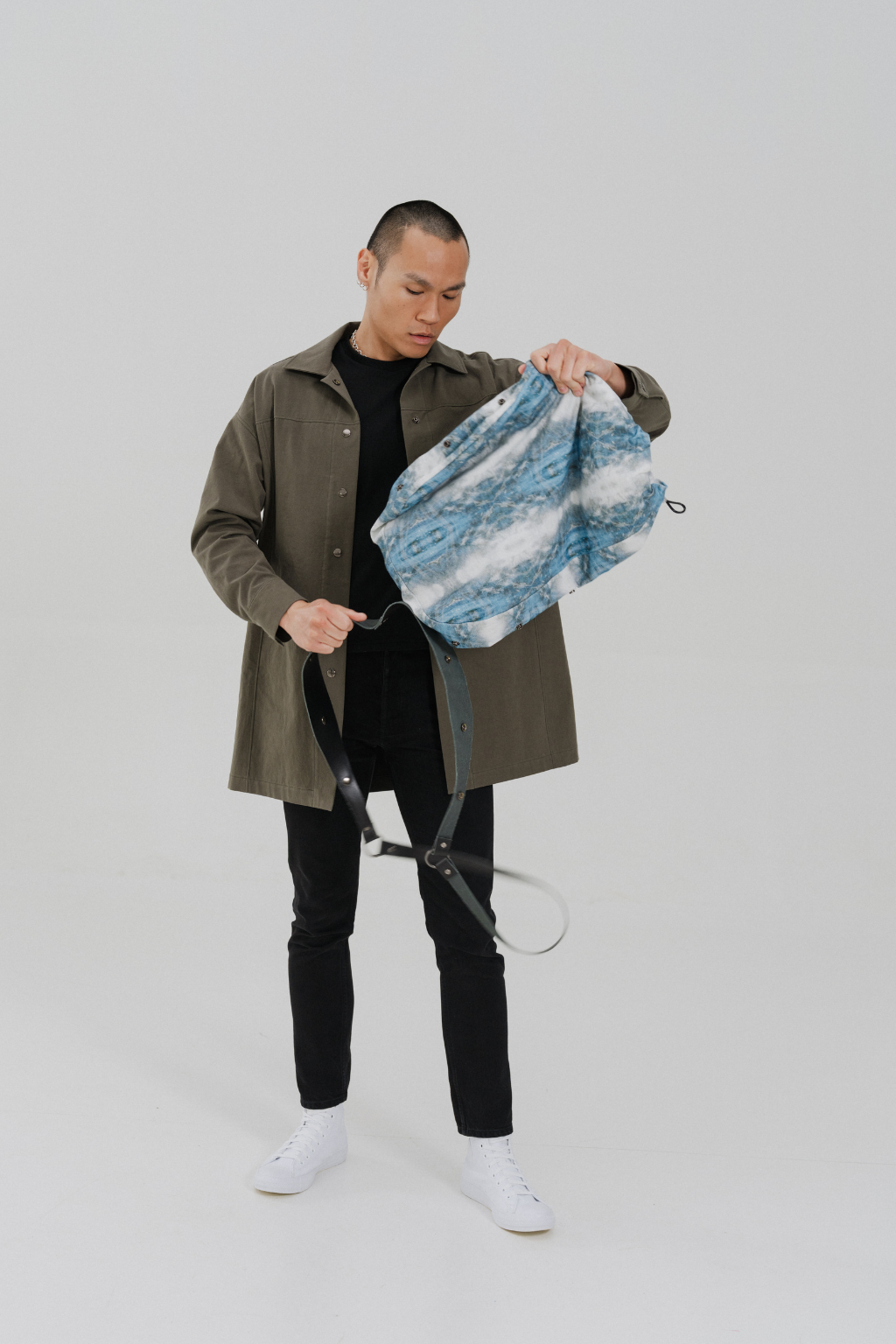 A man wearing an olive green jacket, black shirt, black pants, and white sneakers holds up a blue and white patterned pouch or bag with a black strap against a light gray studio background. Project collab #001: Carryall City Sling bag with collaborative all-over print by September Archive Apparel. Captured by Paper Crane Creative.