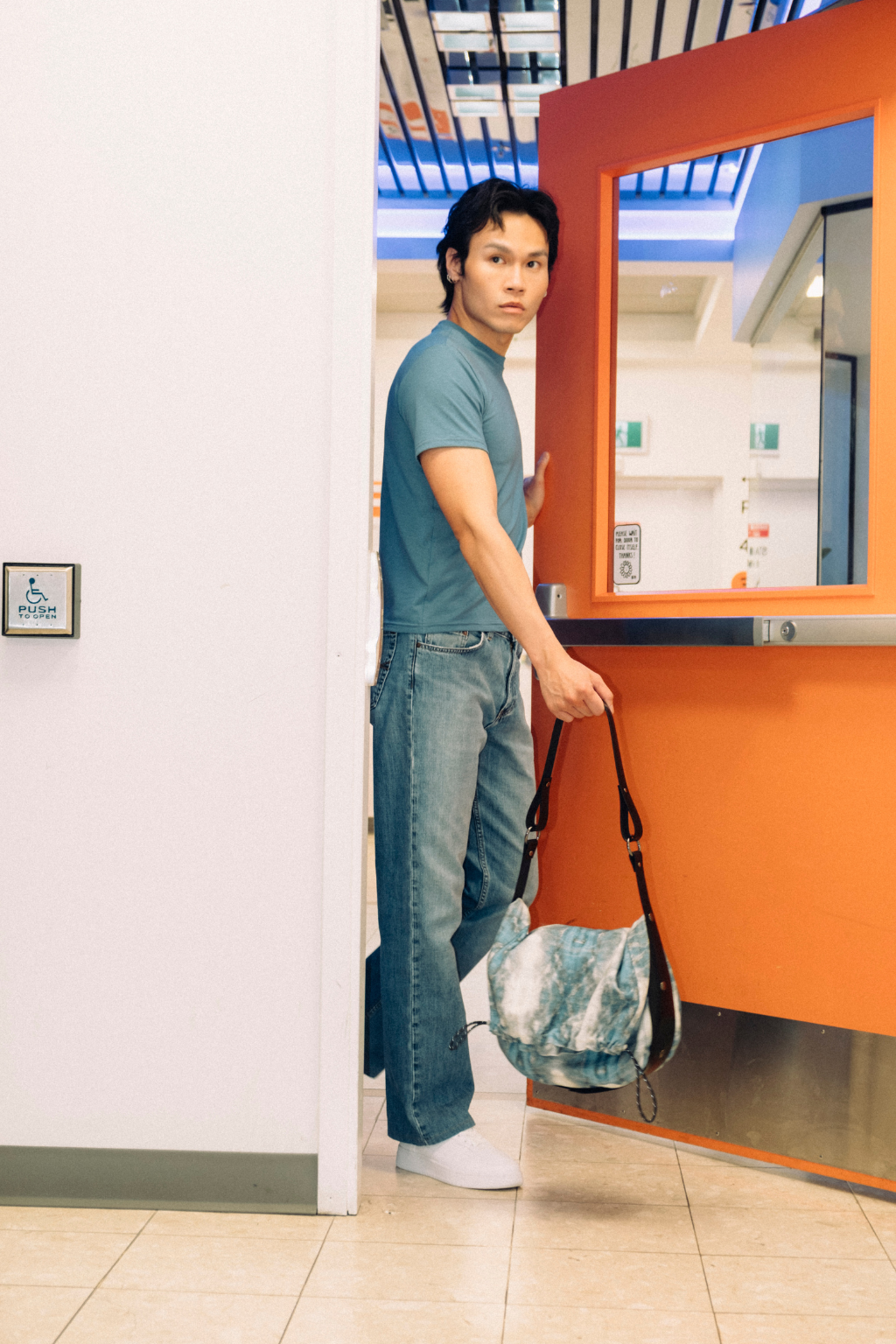 A man wearing a teal t-shirt and blue jeans stands in a doorway holding a blue and white patterned shoulder bag next to a bright orange kiosk in an indoor public space. Project collab #001: Carryall City Sling bag with collaborative all-over print by September Archive Apparel. Captured by Trevor Brady.