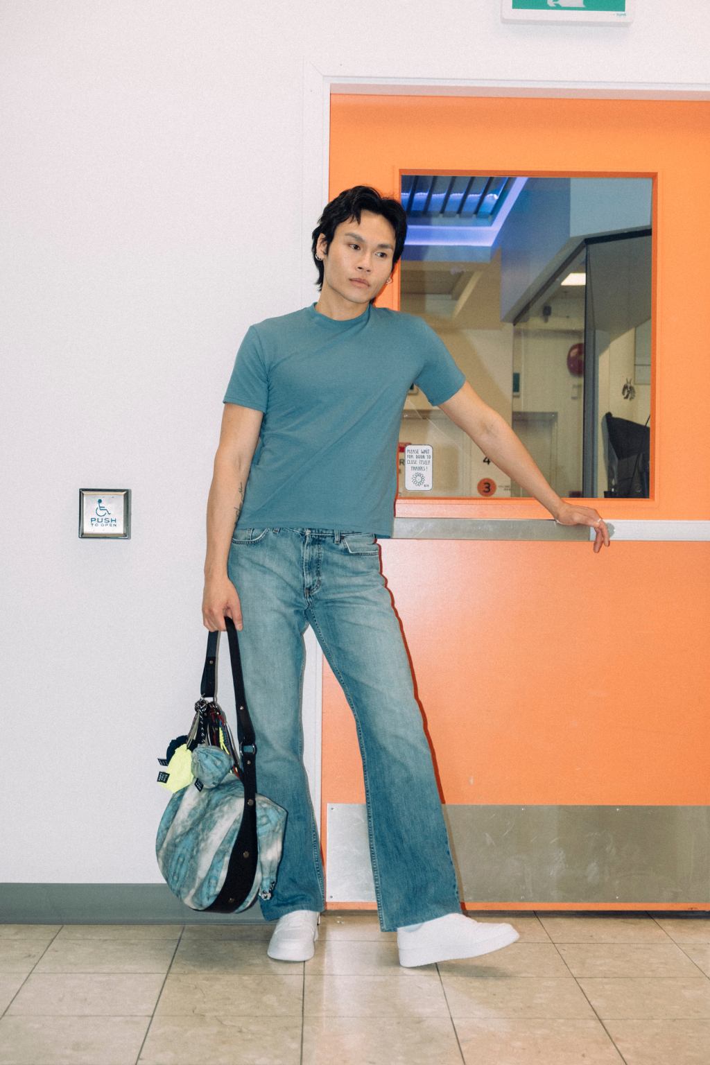 A man wearing a slate blue t-shirt and wide-leg blue jeans stands in a hallway next to an orange service window, holding a blue and white patterned duffel bag. Project collab #001: Carryall City Sling bag with collaborative all-over print by September Archive Apparel. Captured by Trevor Brady.