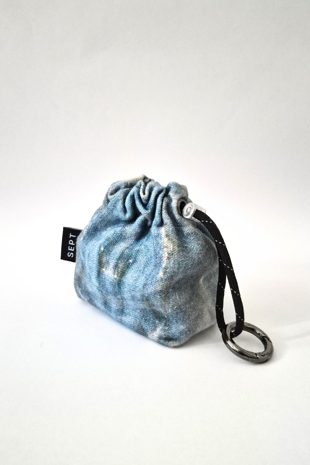 A small drawstring pouch in a washed blue denim-like fabric with a black strap and metal ring, photographed on a plain light background. A small black tag reads SEPT ARC HIVE. Collaboration print 001 mini clip-on pouch by September Archive. Captured by Sheila Wong.