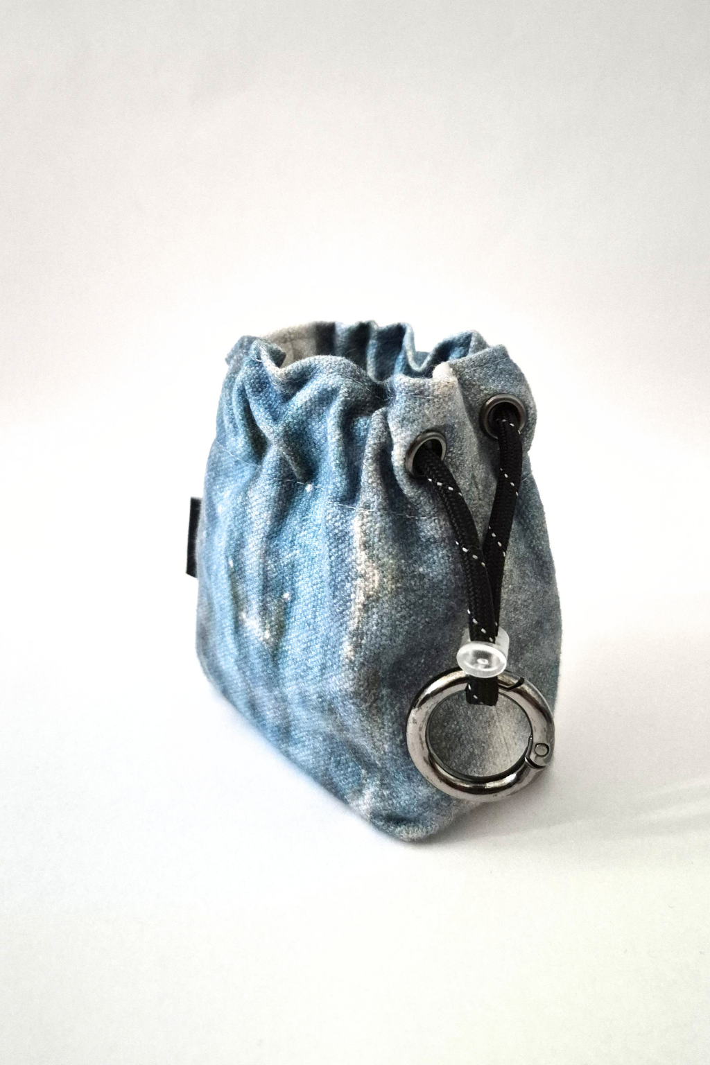 A small blue denim drawstring pouch with black drawstrings and a silver metal ring clasp, photographed on a plain light background. Collaboration print 001 mini clip-on pouch by September Archive. Captured by Sheila Wong.