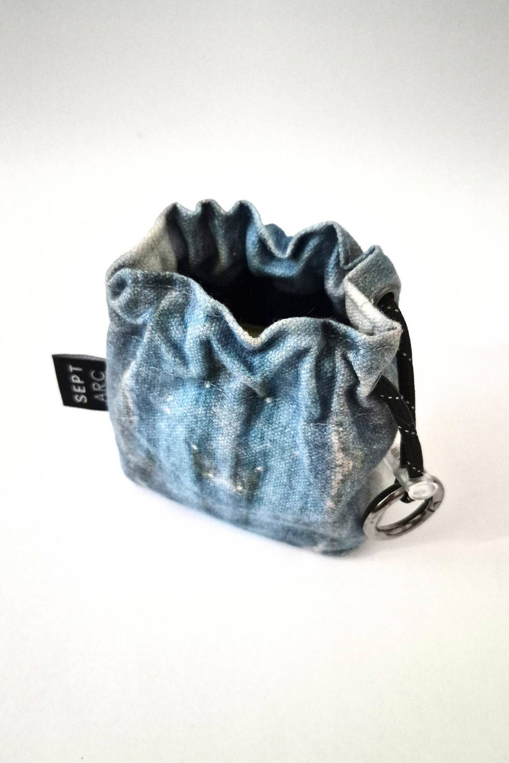 A small denim drawstring pouch in a faded blue wash, featuring a black fabric tag with white text and a silver metal ring attachment, set against a plain white background. Collaboration print 001 mini clip-on pouch by September Archive. Captured by Sheila Wong.