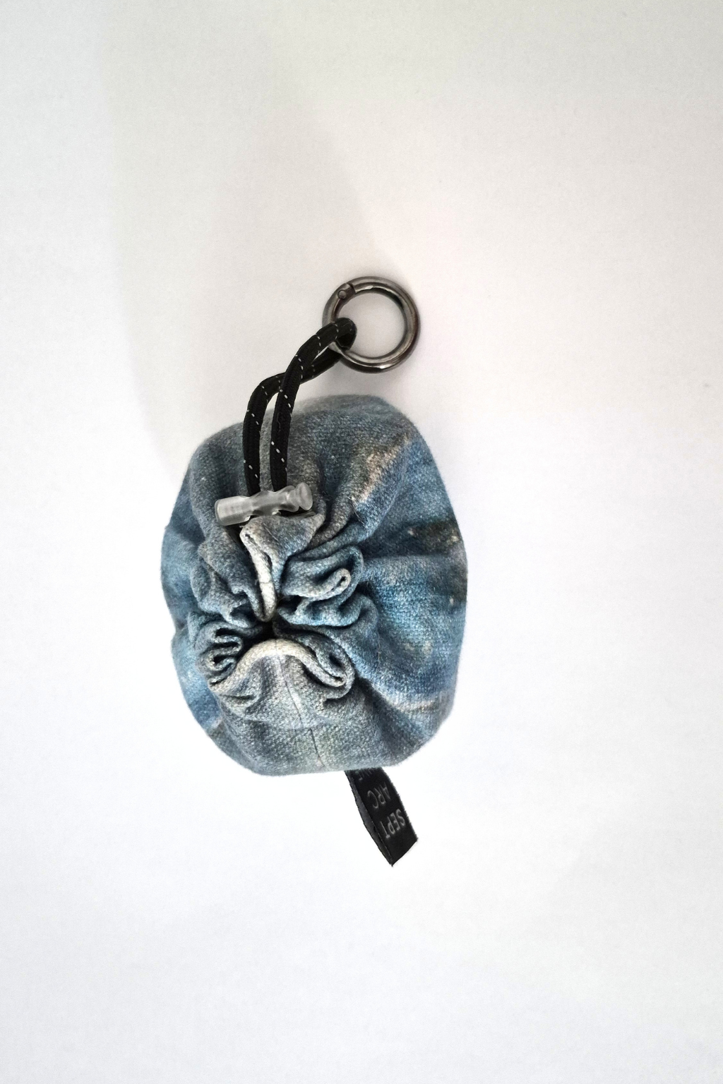 A small blue denim drawstring pouch with a black metal ring and black drawstring, photographed on a plain white background. Collaboration print 001 mini clip-on pouch by September Archive. Captured by Sheila Wong.