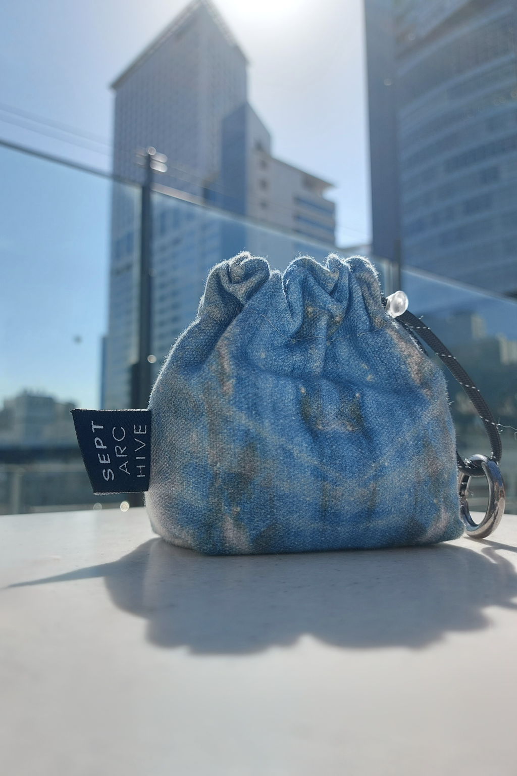 A small drawstring pouch in a blue and white tie-dye patterned canvas, sitting on a white surface with a glass railing and modern buildings in the background. A black fabric tag on the left side reads 'SEPT ARC HIVE'. Collaboration print 001 mini clip-on pouch by September Archive. Captured by Sheila Wong.