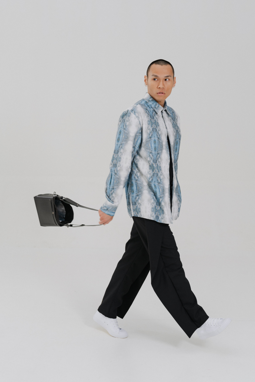 A man walking across a light gray studio background, wearing a blue and white marbled patterned button-up shirt, a black tie, black trousers, white sneakers, and carrying a black shoulder bag. Collaboration print 001 tailored shirt by September Archive. Captured by Paper Crane Creative.