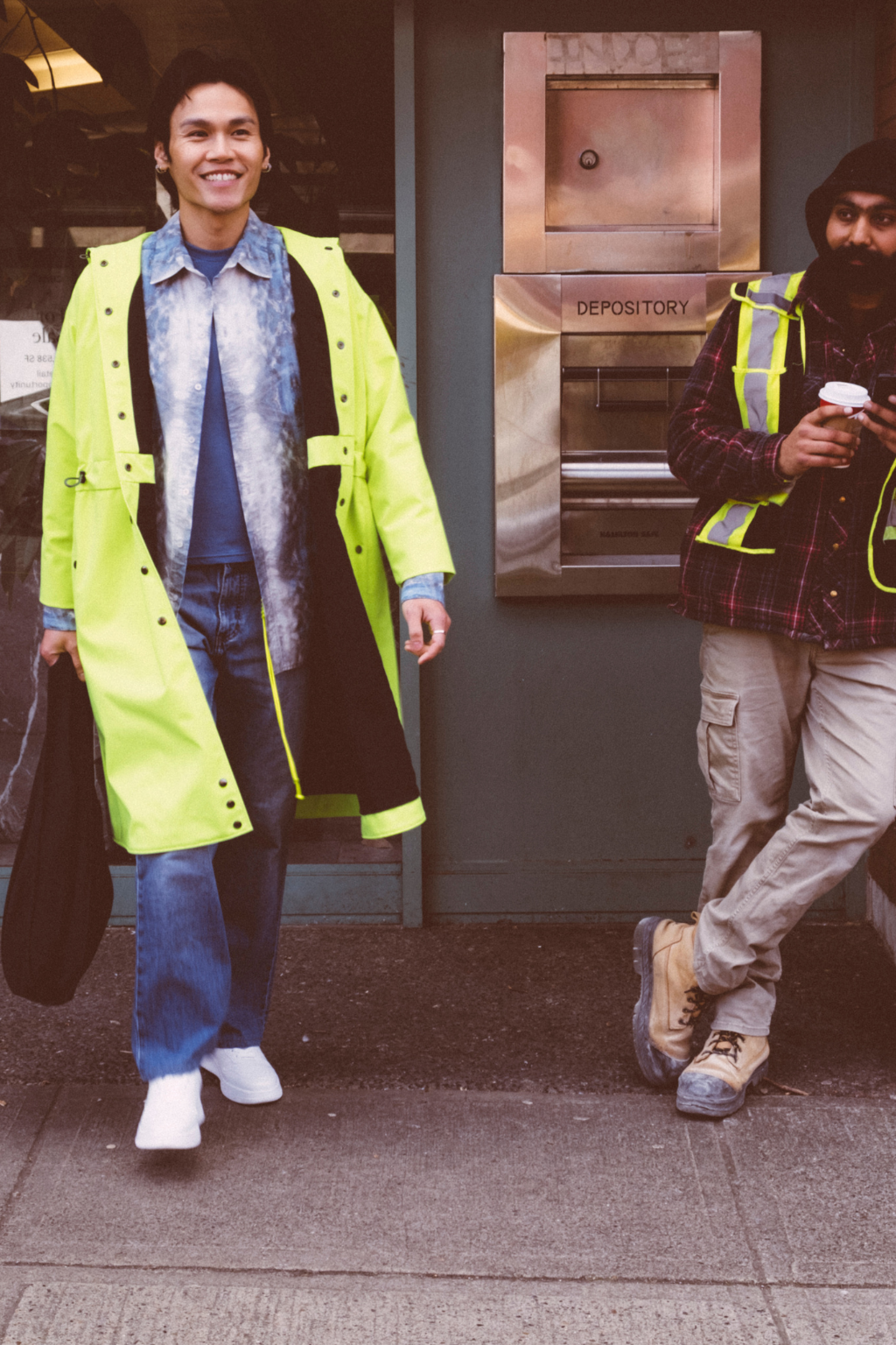 A man wearing a bright neon yellow-green raincoat over a blue shirt and jeans walks outdoors on a sidewalk, while another person in a reflective safety vest stands nearby near a building entrance. Collaboration print 001 tailored shirt by September Archive. Captured by Trevor Brady.