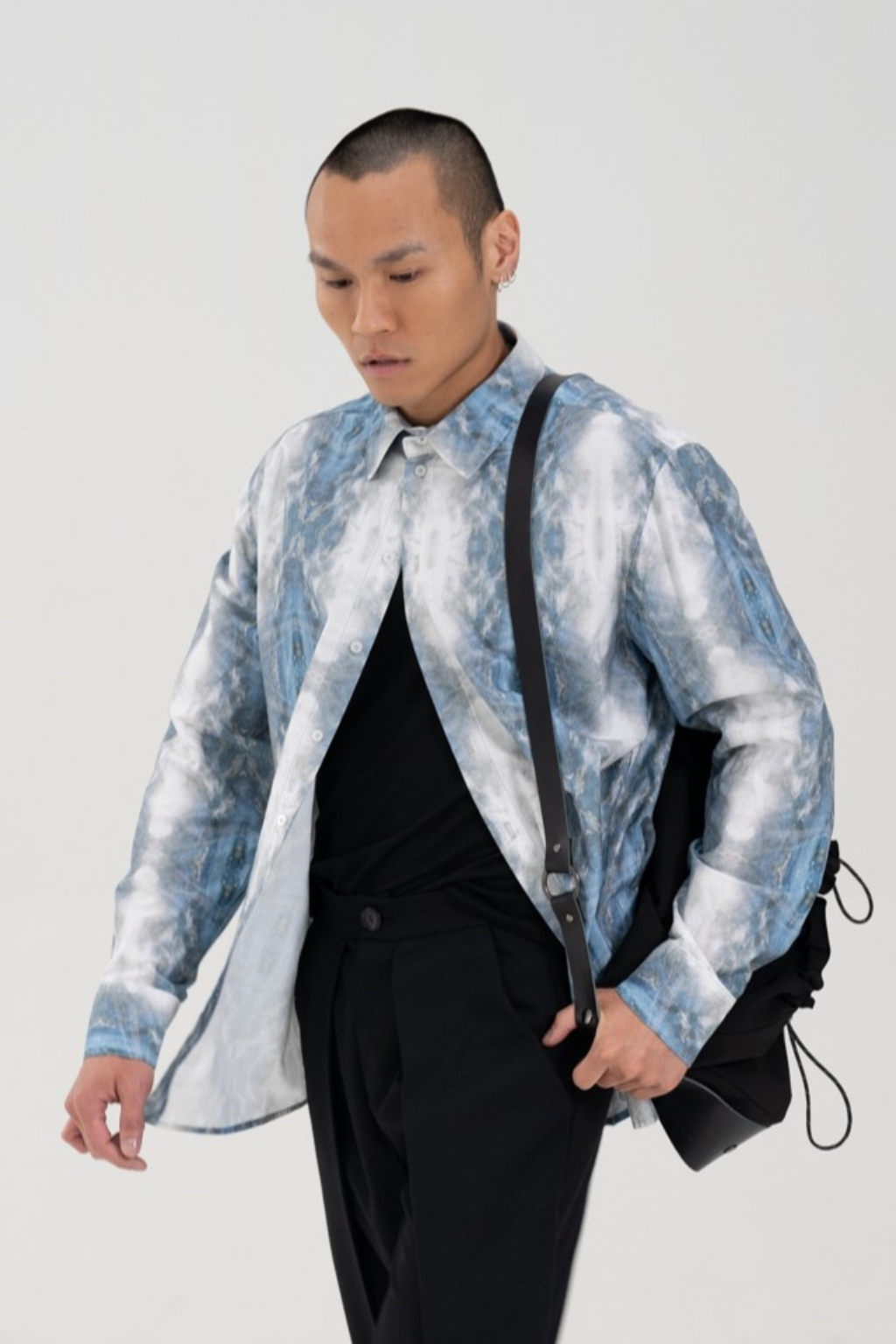 A man wearing a light blue and white marbled patterned overshirt over a black shirt and black trousers, carrying a black shoulder bag against a plain light gray background. Collaboration print 001 tailored shirt by September Archive. Captured by Paper Crane Creative.