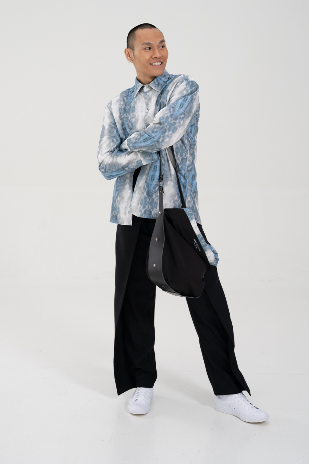 A model stands against a light gray studio background wearing a light blue and white abstract patterned long-sleeve shirt, black wide-leg trousers, white sneakers, and carrying a black shoulder bag. Collaboration print 001 tailored shirt by September Archive. Captured by Paper Crane Creative.