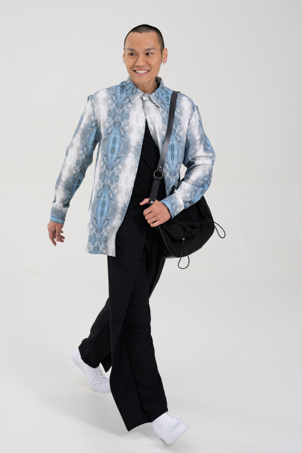 A man modeling a light blue and white patterned long-sleeve shirt jacket over a black shirt and black trousers, carrying a black shoulder bag and wearing white sneakers, against a light gray studio background. Collaboration print 001 tailored shirt by September Archive. Captured by Paper Crane Creative.