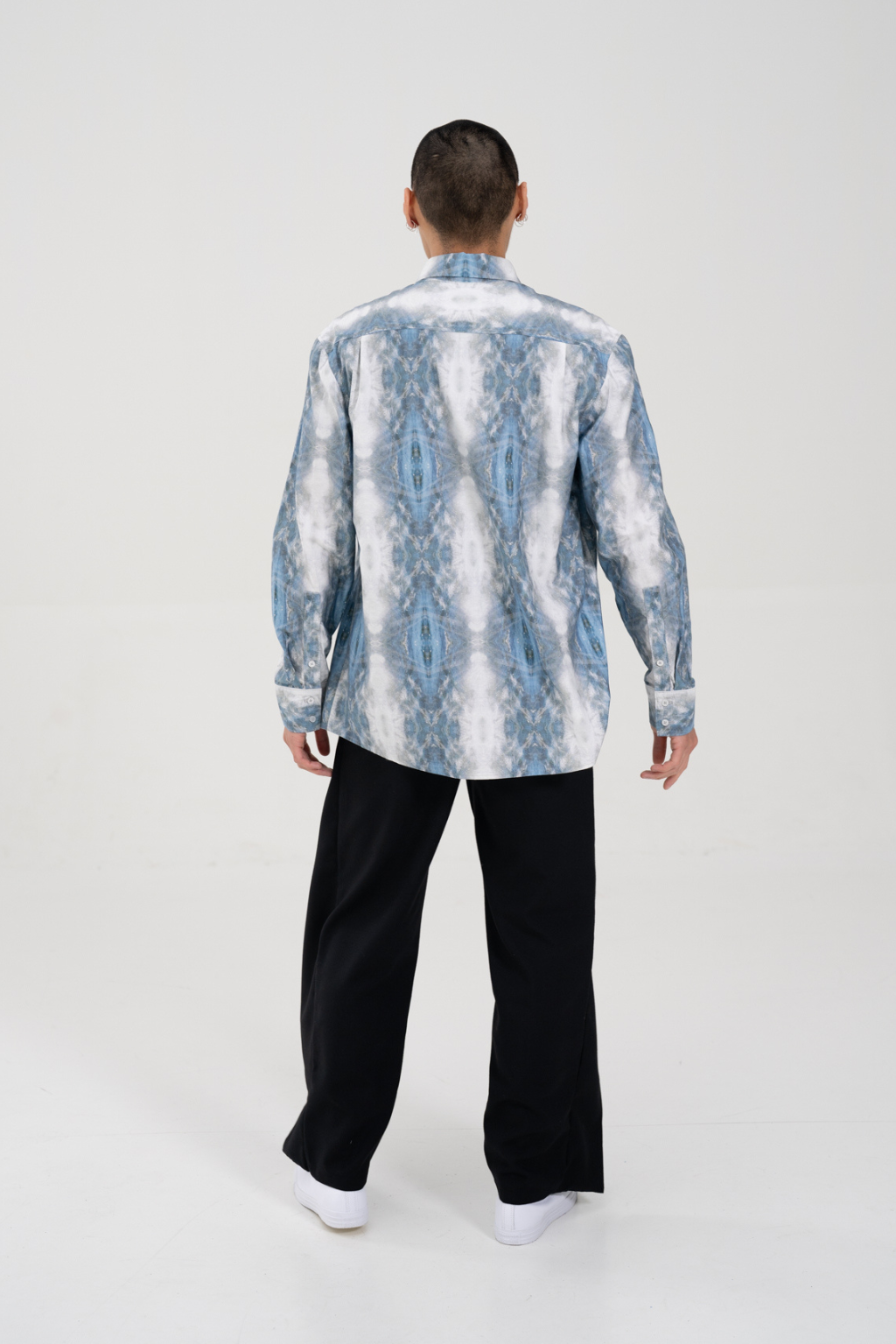 A man viewed from behind wearing a light blue and white patterned button-up shirt and black trousers, standing against a plain white studio background. Collaboration print 001 tailored shirt by September Archive. Captured by Paper Crane Creative.