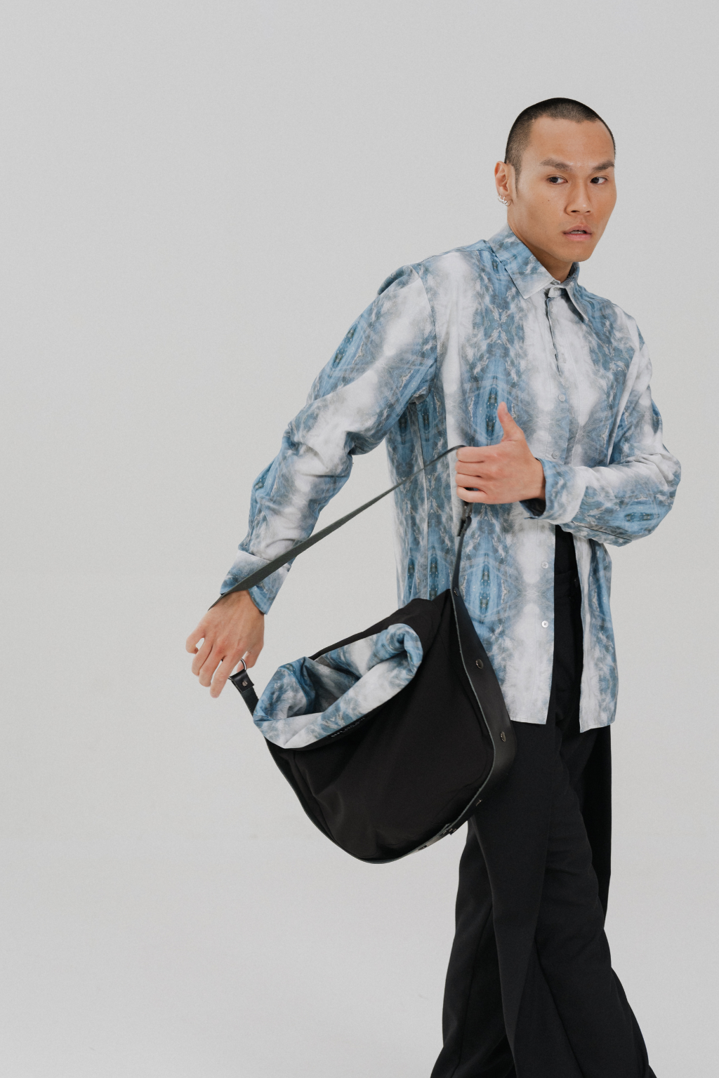A model wearing a blue and white patterned shirt and black trousers carries a black shoulder bag with a blue patterned lining, set against a plain light gray background. Project collab #001: Carryall City Sling bag with collaborative lining print by September Archive Apparel. Captured by Paper Crane Creative.