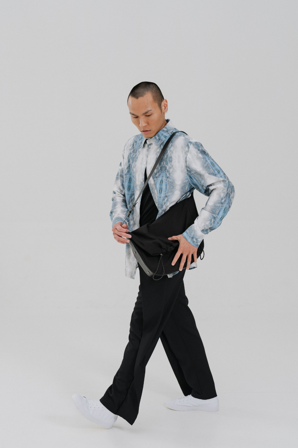 A model walks while wearing a light blue and white marbled long-sleeve shirt, black wide-leg trousers, white sneakers, and carrying a black crossbody bag. Project collab #001: Carryall City Sling bag with collaborative lining print by September Archive Apparel. Captured by Paper Crane Creative.