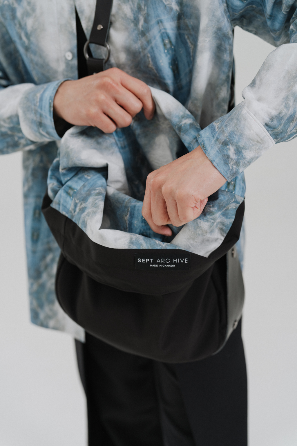 A person wearing a blue and white tie-dye button-up shirt and black pants is shown from the chest down, holding open a black crossbody bag with a white printed logo that reads 'SEPT ARC HIVE'. Project collab #001: Carryall City Sling bag with collaborative lining print by September Archive Apparel. Captured by Paper Crane Creative.