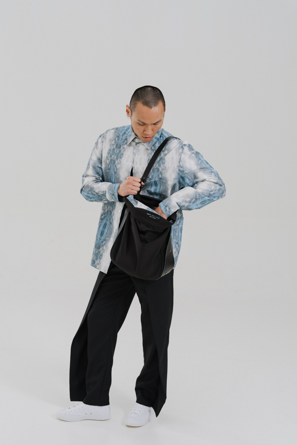 A man wearing a light blue and white patterned button-up shirt, black trousers, and white sneakers is shown from the waist down, looking down into a black shoulder bag he is holding. The background is a clean, neutral light gray studio backdrop. Project collab #001: Carryall City Sling bag with collaborative lining print by September Archive Apparel. Captured by Paper Crane Creative.