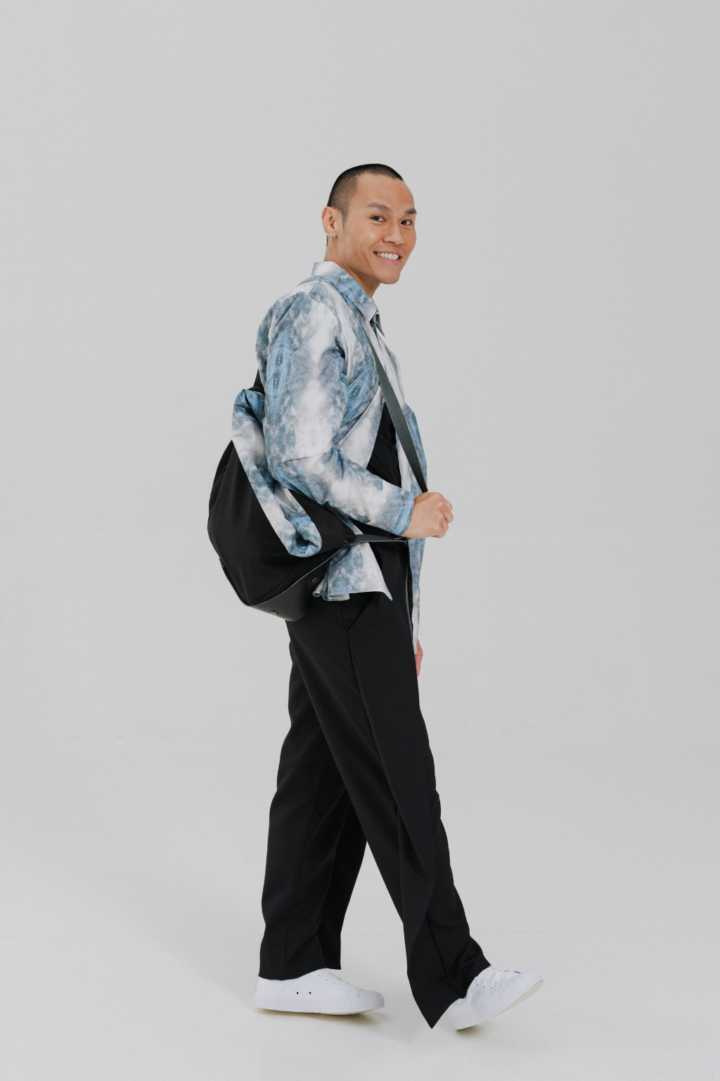 A man wearing a light blue and white tie-dye patterned jacket over a black shirt and black trousers, carrying a black shoulder bag and wearing white sneakers, photographed against a light gray studio background. Project collab #001: Carryall City Sling bag with collaborative lining print by September Archive Apparel. Captured by Paper Crane Creative.