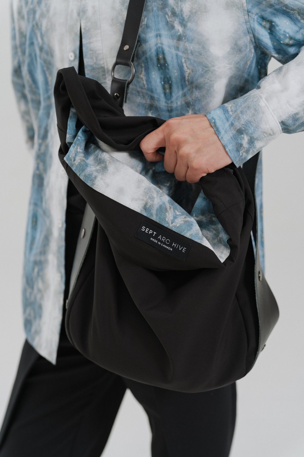 A person wearing a blue and white tie-dye shirt carries a black shoulder bag with a white and blue patterned lining. The brand name 'SEPT ARC HIVE'. Project collab #001: Carryall City Sling bag with collaborative lining print by September Archive Apparel. Captured by Paper Crane Creative.
