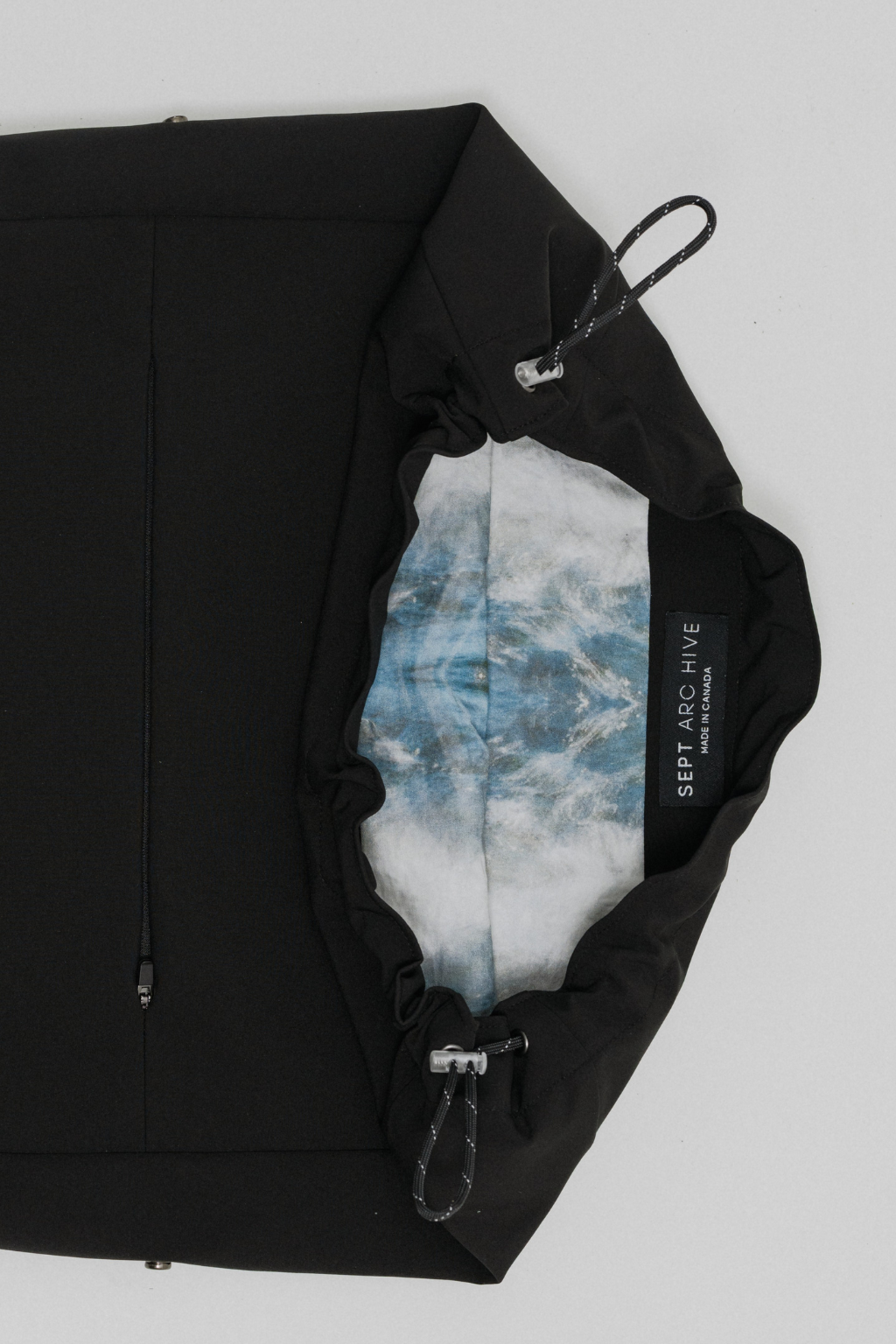 A black zippered pouch or bag shown open on a light gray background, revealing a blue and white marbled interior lining. A black fabric label reads 'SEPT ARCHIVE'. Project collab #001: Carryall City Sling bag with collaborative lining print by September Archive Apparel. Captured by Paper Crane Creative.