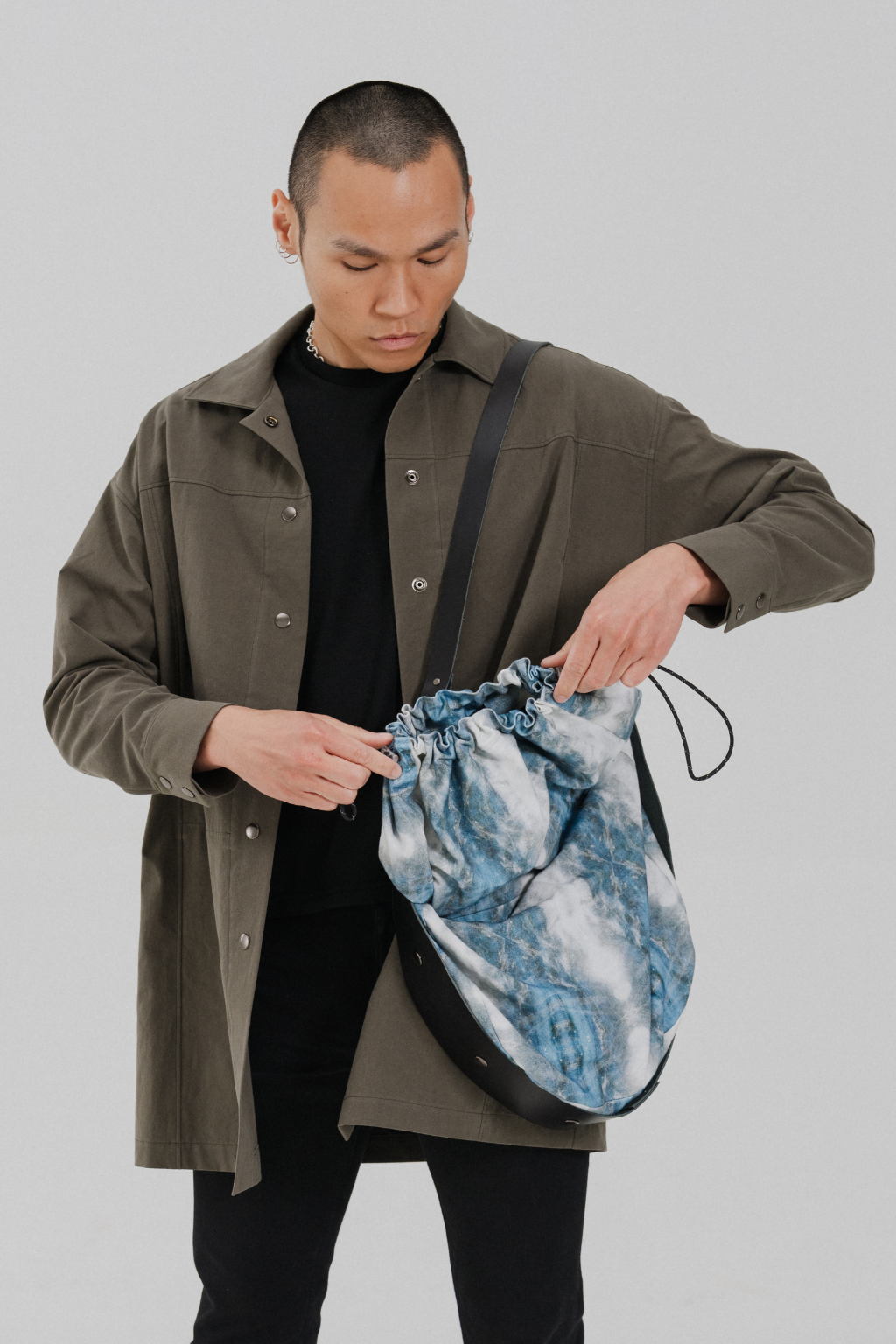 A man wearing an olive green overshirt and black t-shirt is shown from the waist up, demonstrating a blue and white tie-dye drawstring pouch bag with black trim and strap. Project collab #001: Carryall City Sling bag with collaborative all-over print by September Archive Apparel. Captured by Paper Crane Creative.