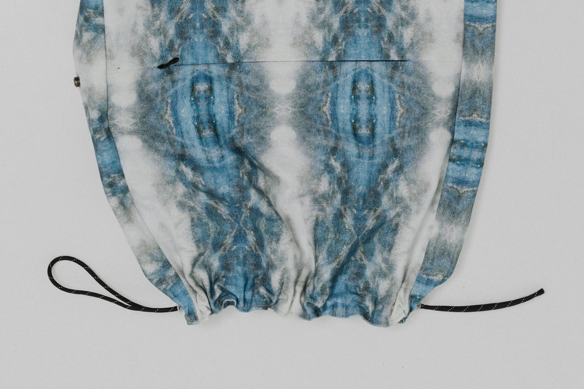 A close-up of a drawstring bag or pouch with a blue and white tie-dye pattern, featuring black drawstrings at the bottom, shot against a light gray background. Project collab #001: Carryall City Sling bag with collaborative all-over print by September Archive Apparel. Captured by Paper Crane Creative.