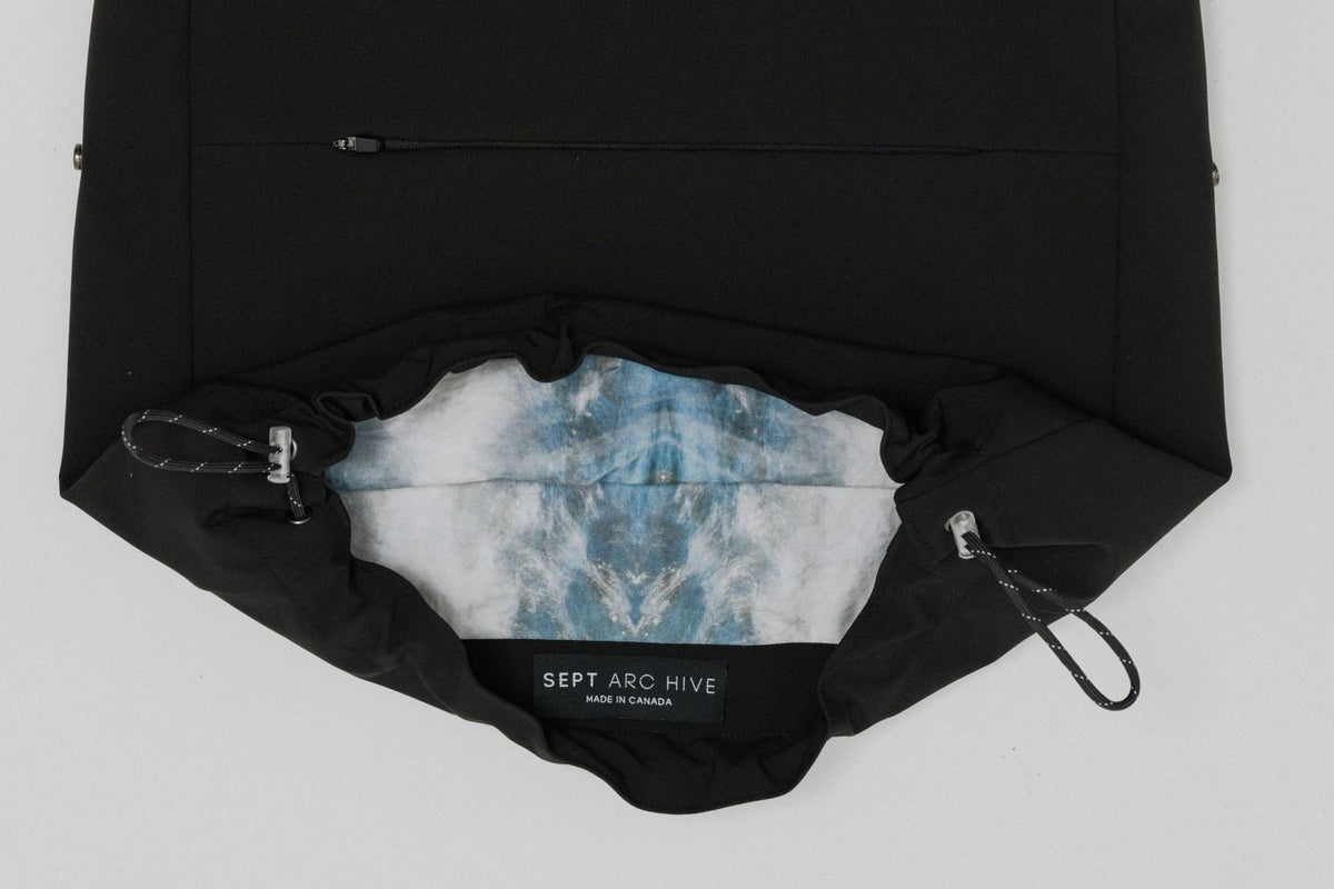 A black drawstring backpack shown open against a light gray background, revealing a blue and white tie-dye patterned face mask inside. A black label at the bottom of the opening reads 'SEPT ARC HIVE'. Project collab #001: Carryall City Sling bag with collaborative lining print by September Archive Apparel. Captured by Paper Crane Creative.