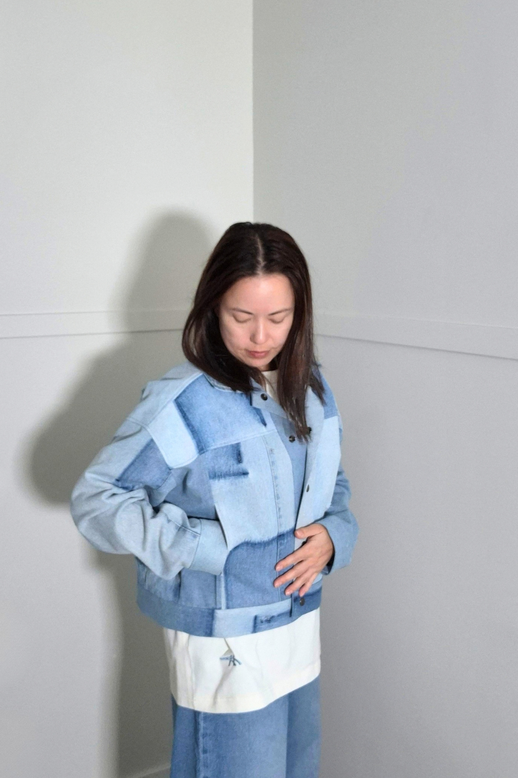 A woman wearing a light blue denim patchwork jacket with a white panel at the hem, standing in a minimalist gray room corner. Upcycled denim jacket by September Archive Apparel. Canadian made denim jacket. One of a kind cropped denim jacket made in Vancouver, Canada. Photographed by Sheila Wong.