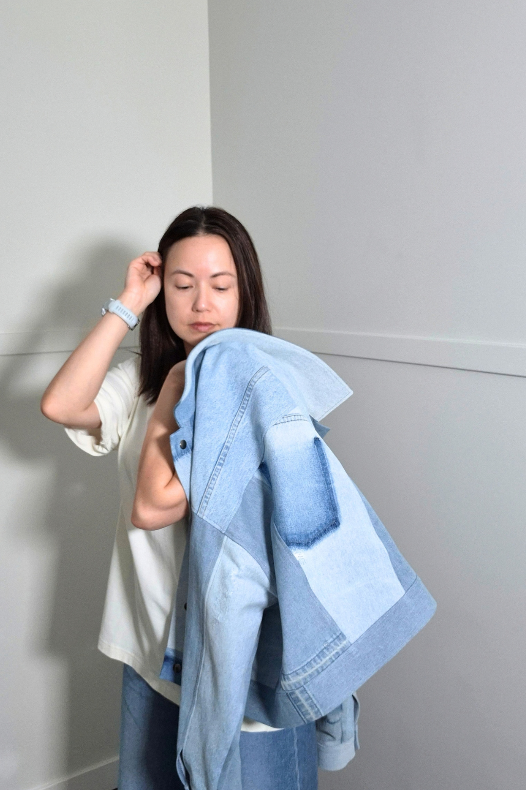 A woman wearing a white t-shirt and blue jeans holds a light blue patchwork denim jacket over her shoulder, standing in a minimalist gray room corner. Upcycled denim jacket by September Archive Apparel. Canadian made denim jacket. One of a kind cropped denim jacket made in Vancouver, Canada. Photographed by Sheila Wong.