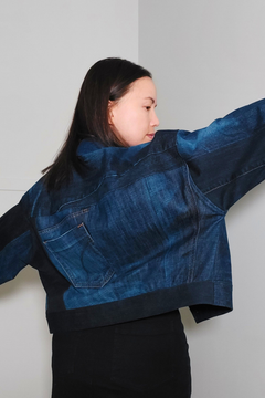 A woman wearing a cropped denim jacket with a blue and black color-blocked design, shown from a side-back angle against a plain light gray wall. Upcycled denim jacket by September Archive. Photographed by Sheila Wong.