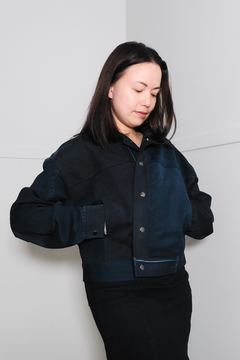 A woman wearing a dark navy blue denim jacket with contrasting suede-like panels on the shoulders and chest, paired with black pants, standing against a plain light gray wall. Upcycled denim jacket by September Archive. Photographed by Sheila Wong.