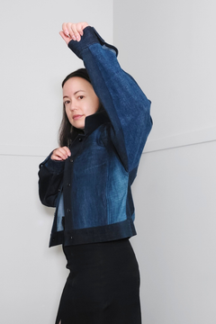 A woman modeling a cropped denim jacket with contrasting dark blue and black panels, posed against a minimalist light gray wall. Upcycled denim jacket by September Archive. Photographed by Sheila Wong.