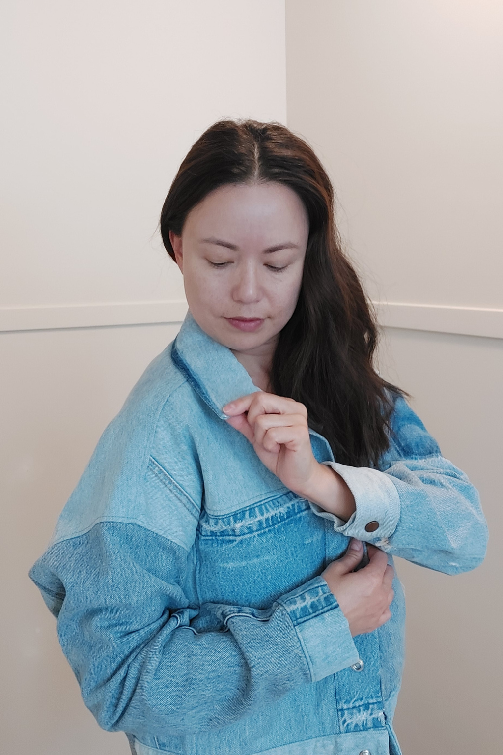 A woman wearing a light blue denim jacket with a color-blocked design, featuring a darker blue front panel and lighter blue denim sleeves and collar, set against a plain beige wall. Upcycled denim jacket by September Archive. Photo by Sheila Wong. 