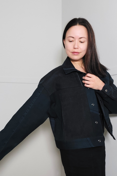 A woman wearing a dark navy blue cropped jacket with contrasting textured panels, posed against a minimalist light gray background. Upcycled denim jacket by September Archive. Photographed by Sheila Wong.