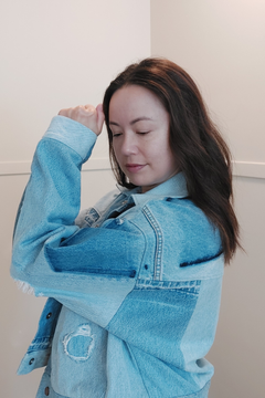 A woman with long brown hair wearing a light blue patchwork denim jacket, posing with her arm raised against a plain off-white wall. Upcycled denim jacket by September Archive. Photo by Sheila Wong. 