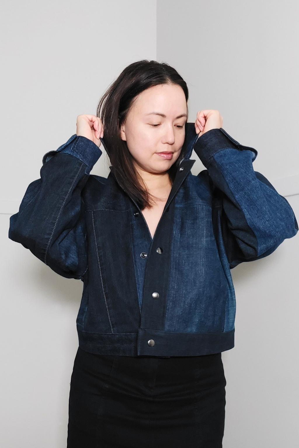 A woman wearing a dark blue denim jacket with contrasting lighter blue denim panels on the shoulders and chest, paired with a black skirt, posing against a plain light gray wall. Upcycled denim jacket by September Archive. Photographed by Sheila Wong.