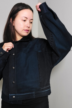 A woman wearing a dark indigo denim jacket with a button-up front and wide sleeves, holding up one sleeve to show the fabric texture and length. Upcycled denim jacket by September Archive. Photographed by Sheila Wong.