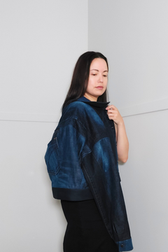 A woman wearing a dark blue indigo denim jacket with a high collar and oversized silhouette, paired with a black skirt, standing against a minimalist white wall. Upcycled denim jacket by September Archive. Photographed by Sheila Wong.