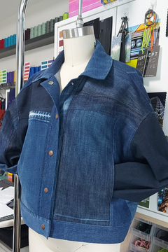 A cropped denim jacket displayed on a mannequin torso in a retail or studio setting. The jacket has a medium blue washed front panel, dark navy sleeves and collar, and a small white embroidered detail on the left chest.
