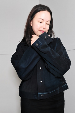 A woman wearing a dark navy blue denim jacket with silver snap buttons, posing against a light gray wall. Upcycled denim jacket by September Archive. Photographed by Sheila Wong.
