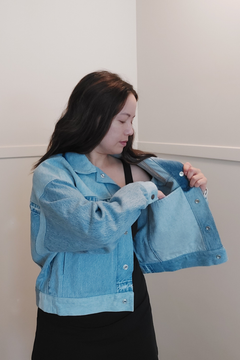 A woman wearing a light blue cropped denim jacket over a black dress, holding the jacket open to show the pockets and lining. Upcycled denim jacket by September Archive. Photo by Sheila Wong. 