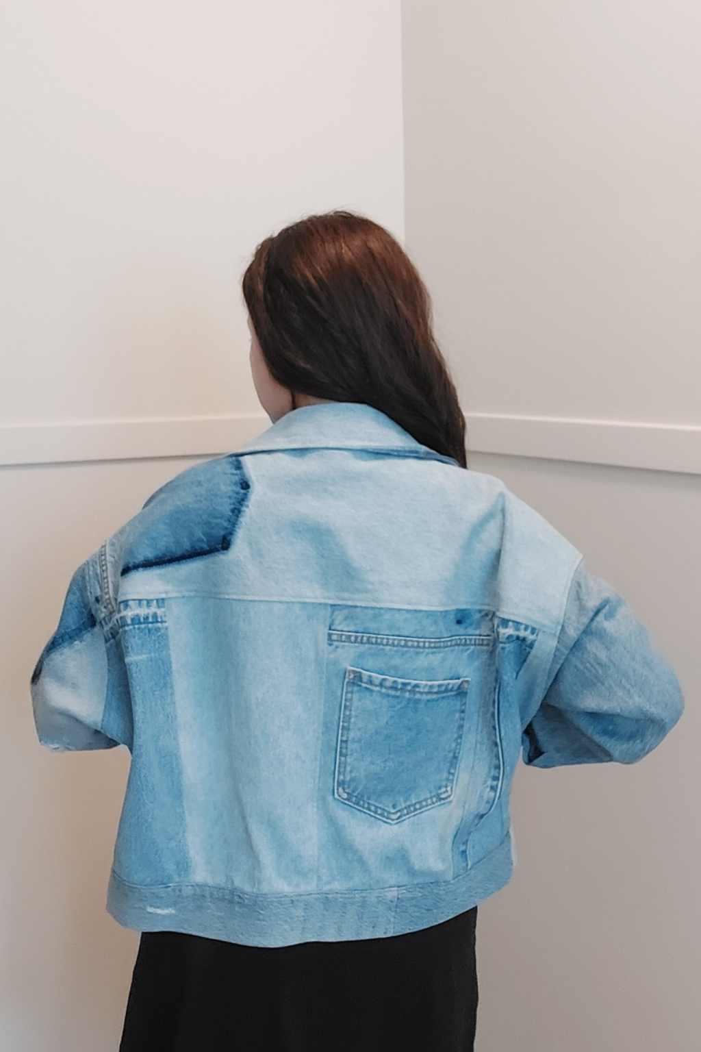 A person viewed from behind wearing a light blue denim jacket with a patchwork back design and a single back pocket, paired with a black skirt, against a plain white wall. Upcycled denim jacket by September Archive. Photo by Sheila Wong. 