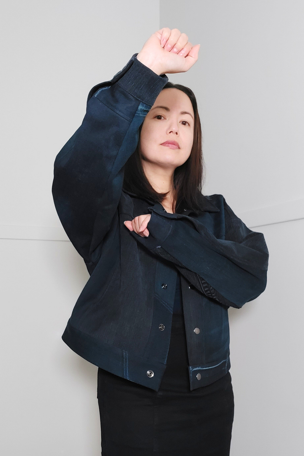 A woman wearing a cropped dark navy denim jacket over a black top and black skirt, posing with one arm raised against a light gray background. 